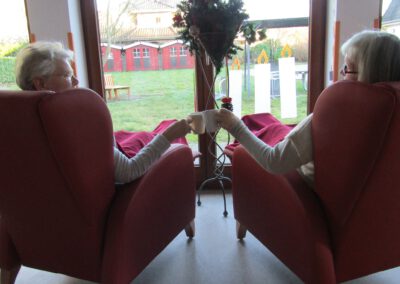 Zwei Bewohnerinnen sitzen sich in roten Sesseln gegenüber, stoßen mit Tassen an und genießen die weihnachtliche Stimmung. Zwischen ihnen steht ein festlich geschmückter Adventskranz vor einem großen Fenster mit Blick ins Grüne. Über ihnen hängen Sterne und Tannenzapfen als Dekoration.
